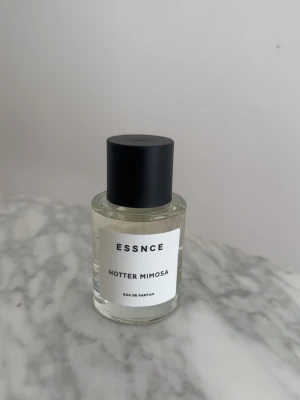 ESSNCE Hotter Mimosa Eau de Parfum - ESSNCE Hotter Mimosa Eau de Parfum kommer i en stilren, transparent glasflaska med svart cylinderformad kork. Doften har en modern och minimalistisk design med vit etikett och tydlig text. Perfekt för dig som gillar fräscha och unika parfymer.