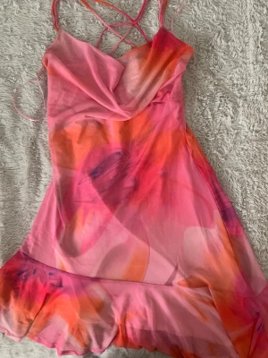 Rosa och orange kortklänning med volang - Säljer en färgstark kortklänning med tunna axelband och snygg omlott-drapering fram från Behrska. Klänningen har ett abstrakt mönster i rosa, orange och lila toner samt volangkant nertill. Materialet känns som lätt polyester, perfekt för varma dagar. Axelbanden är korsade i ryggen för extra detalj. Perfekt för utomlands!