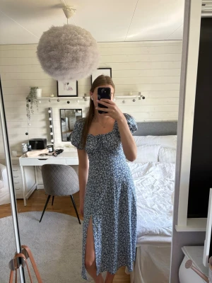 Blommig blå långklänning med slits - Söt blå långklänning med vitt blommönster och puffärmar. Klänningen har en smickrande v-ringning, slits framtill och öppen rygg där dragkedja finns. Perfekt för sommarens alla tillfällen. Tillverkad i ett lätt och luftigt material.
