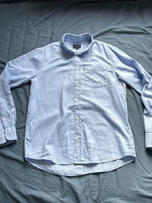 Ljusblå skjorta från Lexington - Klassisk ljusblå skjorta från Lexington med button-down krage och bröstficka. Tillverkad i bomullstyg som känns mjukt och svalt. Perfekt till jeans eller chinos för en clean och stilren look.