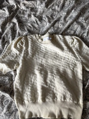 Vit quiltad tröja med puffärm - Säljer en vit quiltad tröja med korta puffärmar och rund hals. Tröjan har ett diskret rutmönster i tyget och breda ribbade muddar vid ärmslut och nederkant. Perfekt för dig som gillar en stilren men ändå trendig look.