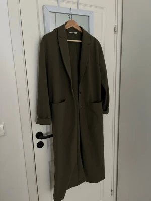 Olivgrön lång kappa från ONLY - Stilren olivgrön kappa från ONLY i rak modell med klassisk krage och två stora fickor framtill. Kappan har långa ärmar och stängs med knappar. Perfekt för dig som gillar minimalistisk och trendig look.