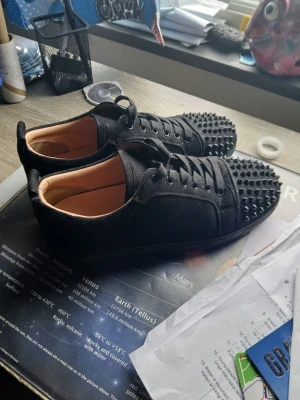 Svarta sneakers med nitar - Säljer ett par svarta sneakers med coola svarta nitar på tån och ovandelen. Skorna har en låg profil, snörning och är gjorda i mockaliknande material med beige innerfoder. Perfekta för dig som vill sticka ut med din stil. Det saknar en tag på skorna men har två extra i en påse.