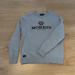 Grå stickad tröja från Morris Gents - Snygg grå stickad tröja från Morris Gents med broderad logga och brittisk flagga på bröstet. Tröjan har rund hals, ribbade muddar och nederkant. Perfekt för en stilren och avslappnad look.