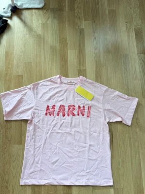 Ljusrosa Marni t-shirt med tryck - Säljer en ljusrosa t-shirt från Marni med stort rött logotyptryck på bröstet. T-shirten har klassisk rund hals och korta ärmar. Tillverkad i mjuk bomull för en skön känsla. Perfekt för dig som gillar stilrena plagg med en twist.