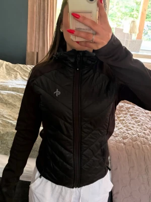 Svart vindjacka från Peak Performance - Snygg svart vindjacka från Peak Performance med dragkedja framtill och huva. Jackan har ett diskret logotryck på bröstet och quiltade partier på framsidan. Perfekt för blåsiga dagar och enkel att matcha med olika outfits.