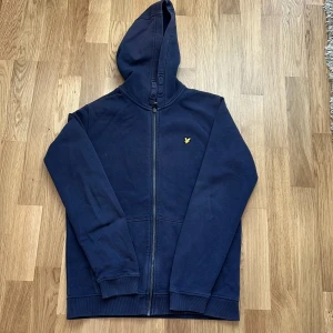 Marinblå hoodie från Lyle & Scott - Marinblå hoodie från Lyle & Scott med dragkedja och klassisk gul logga på bröstet. Tröjan har huva, långa ärmar och är gjord i mjuk bomull. Perfekt för chill dagar eller när du vill ha en enkel och stilren look.