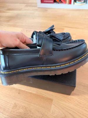 Svarta loafers från Dr. Martens - Snygga svarta loafers från Dr. Martens i läder med klassisk tofs och markerad gul söm runt sulan. Skorna har en chunky sula och en stilren design som passar perfekt till jeans eller kostymbyxor.
