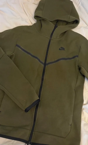 Olivgröna Nike Tech Fleece mjukisbyxor - Säljer ett par olivgröna Nike Tech Fleece mjukisbyxor med svarta detaljer och dragsko i midjan. Byxorna har smal passform, muddar vid bensluten och en stor dragkedjeförsedd ficka på sidan. Perfekta för chill eller träning.