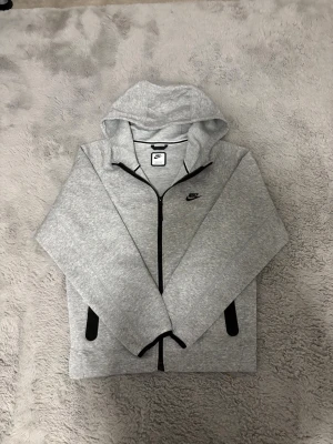 Grå zip hoodie från Nike - Snygg grå hoodie från Nike med dragkedja framtill och svart Nike-logga på bröstet. Hoodien har huva och svarta detaljer vid fickorna. Tillverkad i mjukt sweatshirtmaterial som är perfekt för en avslappnad stil.