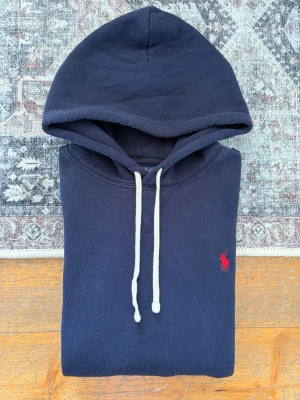 Marinblå Polo Ralph Lauren Hoodie - Storlek M - Hej! 👋 Säljer en klassisk marinblå Polo Ralph Lauren hoodie i storlek M med röd pony-logga. 🐎✨ Tröjan är i riktigt fint skick, välvårdad och helt utan anmärkningar. Personen på bilden är ca 177 cm och den upplevs som något stor – passar troligtvis allra bäst på någon runt 180–185 cm. Självklart 100% äkta med originaltvättlappar kvar (ingen QR-kod på just denna årsmodell). Ett tidlöst premium-basplagg! Fraktar snabbt! 🚛📦💬 