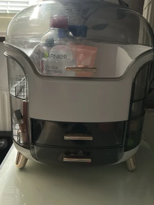 Smink- och hudvårdsförvaringsbox - Praktisk förvaringsbox för smink och hudvårdsprodukter i vit och grå plast med transparent lock och guldfärgade handtag. Boxen har flera fack och lådor för enkel organisering av dina skönhetsprodukter. Perfekt att ha på sminkbordet eller i badrummet.