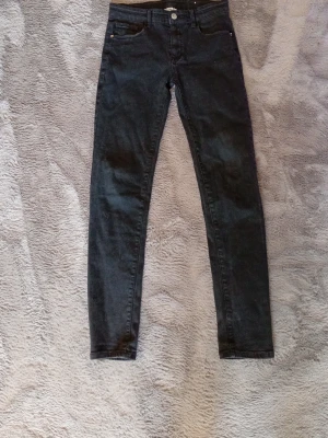 Jeans - Säljer ett par svarta  jeans med hög midja och klassisk femficksdesign. Jeansen har smal passform hela vägen ner och stängs med knapp och dragkedja. Perfekta för dig som gillar en clean och enkel look.