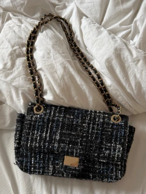Cool tweed tiamo handväska - Stilren svart handväska i tweed med inslag av blått och vitt. Väskan har en guldfärgad kedjerem och matchande guldfärgad platta framtill. Väskan är från tiamo!