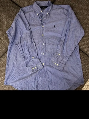 Ralph Lauren Oxford Skjorta - Klassisk blå skjorta från Ralph Lauren med button-down krage och broderad logga på bröstet. Skjortan har långa ärmar, vita knappar och är tillverkad i bomull. Perfekt till en avslappnad men ändå stilren look.