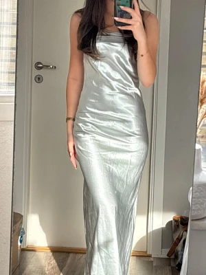 Silverfärgad blå långklänning i satin - Elegant silverfärgad blå  långklänning i skimrande satinmaterial. Klänningen har en slimmad passform och är ärmlös med raka axelband. 