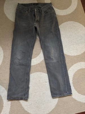 Grå raka jeans från Nudie Jeans - Snygga grå jeans från Nudie Jeans med rak passform och klassisk femficksdesign. Jeansen har subtila slitningar och är tillverkade i mjuk denim. Perfekta för en avslappnad och trendig stil. W32 W32