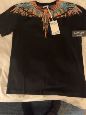 Svart t-shirt Marcelo Burlon - Svart t-shirt från Marcelo Burlon County of Milan med färgstarkt grafiskt mönster i orange, blått och turkost runt halsen. Klassisk passform och rund hals. Perfekt för dig som vill sticka ut med en unik design.