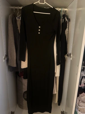 Svart långklänning med knappar - Svart långklänning med v-ringning och tre dekorativa knappar framtill. Storlek S Ej använd. Har 2 stycken!