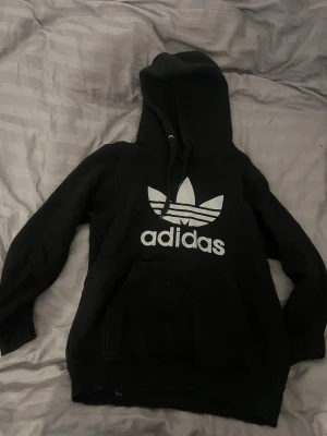 Svart Adidas hoodie med stor logga - Svart hoodie från Adidas med stor vit Trefoil-logga och text på bröstet. Klassisk känguruficka framtill och justerbar huva med snören. Tillverkad i mjukt bomullsmaterial, perfekt för en avslappnad streetwear-look.