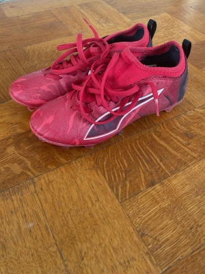 Röda puma fotbollsskor - Säljer ett par röda puma fotbollsskor med svart och vit detalj på sidan med svart puma-logga. Skorna har snörning och mönstrad ovandel i syntetmaterial för bra grepp och känsla på planen.