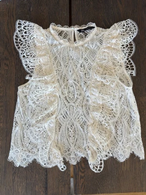 Vit spetsblus med volang från Zara S - Supersöt vit spetsblus från Zara med detaljerade broderier och volanger längs axlarna. Blusen är ärmlös och har en rund halsringning samt knapp i nacken. Perfekt för dig som gillar romantisk och bohemisk stil.