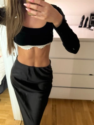 Svart croppad topp med spetskant - Trendig svart croppad topp med lång ärm och vit spetskant nedtill. Toppen har en tight passform och är perfekt för dig som vill ha en edgy och stilren look. Materialet känns mjukt och stretchigt, vilket gör den bekväm att bära.