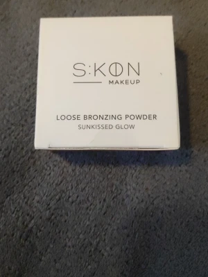 SKON Loose Bronzing Powder - Loose Bronzing Powder från SKON Makeup i nyansen Sunkissed Glow. Kommer i en stilren vit förpackning. Produkten är ett löspuder som ger en solkysst finish och naturlig lyster till huden. Perfekt för att framhäva ansiktets konturer och skapa glow.