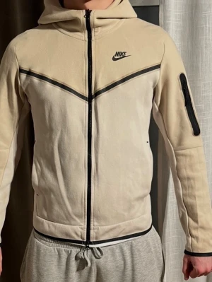 Beige Nike tech fleece - Snygg beige hoodie från Nike med svart dragkedja och logga på bröstet. Hoodien har en huva, svarta detaljer längs bröstet och en praktisk ficka med dragkedja på ena ärmen. Tillverkad i mjukt material som känns skönt mot huden.