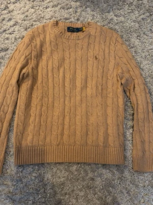 Beige kabelstickad tröja Polo Ralph Lauren - Säljer en beige kabelstickad tröja från Polo Ralph Lauren med rund halsringning och diskret broderad logga på bröstet. Tröjan har långa ärmar och ribbade muddar vid ärmslut och nederkant. Perfekt för dig som gillar klassisk och stilren look.