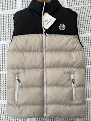 Beige och svart dunväst Moncler - Snygg dunväst från Moncler i beige och svart med hög krage och två dragkedjeförsedda fickor framtill. Västen har en quiltad design och Moncler-logga på bröstet. Perfekt för lager-på-lager och håller dig varm under kyliga dagar.