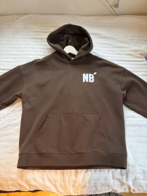 Brun hoodie från No Biggie - Säljer en brun hoodie från No Biggie med vit broderad text 'NB' fram och 'NO BIGGIE' stort på ryggen. Hoodien har en klassisk känguruficka och huva. Perfekt för dig som gillar streetwear och vill ha en chill look.