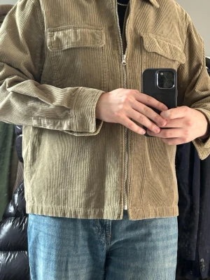 Beige manchester overshirt med dragkedja - Stilren beige overshirt i manchestertyg med dragkedja framtill och två bröstfickor med lock. Rak passform och klassisk krage, perfekt att bära över en t-shirt. Ger en avslappnad men ändå snygg look.