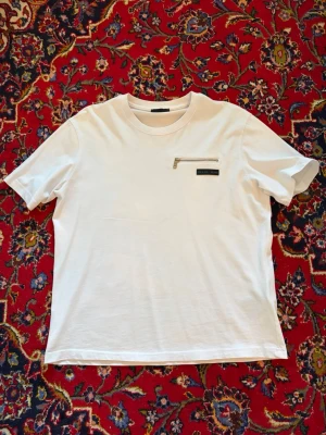 Vit t-shirt från Prada med dragkedja - Vit t-shirt från Prada med rund hals och cool detalj: en liten ficka med silverfärgad dragkedja på bröstet samt Prada Milano-logga. Materialet är mjukt och känns lyxigt, perfekt för dig som gillar stilrena designer.