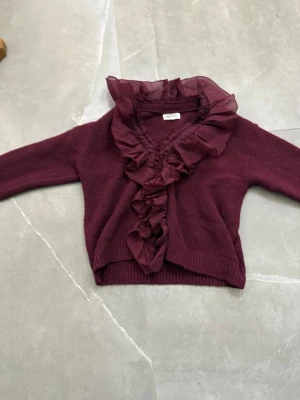 Vinröd kofta med volang från Name It - Supersöt vinröd kofta från Name It med stora volanger i chiffong längs kragen och framtill. Koftan har långa ärmar och ribbade muddar vid ärmslut och nederkant. Perfekt för dig som gillar en romantisk och trendig stil.