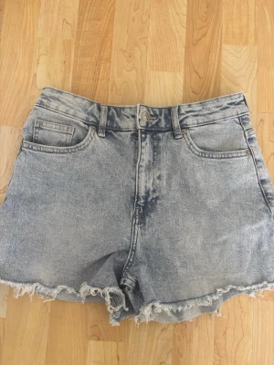 Ljusblå denimshorts med fransig kant - Säljer ett par ljusblå denimshorts med hög midja och rå, fransig kant. Klassisk femficksmodell med knapp och dragkedja framtill. Perfekta för sommaren och ger en avslappnad vibe.