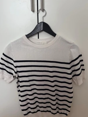 Vit och svart randig topp H&M - Kortärmad topp från H&M i vit med svarta horisontella ränder. Klassisk rund halsringning och ribbade kanter. Perfekt för en enkel och stilren look. Materialet känns mjukt och lätt, troligen bomull eller bomullsblandning.