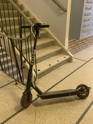 Svart elsparkcykel med röda detaljer - Säljer en svart elsparkcykel med röda detaljer på hjulen och gula stjärnklistermärken på styret. Elscootern har en robust ram i metall och en vit laddkabel virad runt styret. Perfekt för att ta sig runt i stan på ett smidigt sätt.