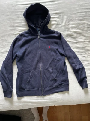 Mörkblå hoodie från Polo Ralph Lauren - Mörkblå hoodie från Polo Ralph Lauren med dragkedja framtill och klassisk röd logga broderad på bröstet. Hoodien har fickor och en stor huva. Tillverkad i mjukt bomullsmaterial som är perfekt för chill dagar. Jag är 177 cm lång och passar mig bra!