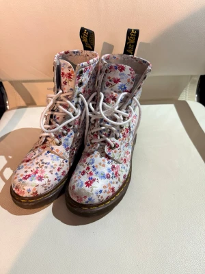 Blommiga Dr. Martens stövletter - Säljer ett par unika Dr. Martens stövletter med vit bas och färgglatt blommönster i rött, blått och lila. Klassisk gul söm runt sulan, vita snören och svarta dragflikar med gul text. Tillverkade i slitstarkt läder och har den ikoniska grova sulan.