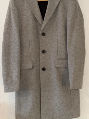 Grå ullkappa från Studio Total - Stilren grå kappa från Studio Total. Klassisk modell med tre knappar framtill. Perfekt för dig som gillar minimalistisk och tidlös stil.