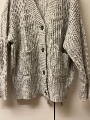 Grå stickad kofta med fickor - Mysig grå stickad kofta med v-ringning och stora knappar framtill. Koftan har två praktiska fickor och ribbade muddar vid ärmslut och nederkant. Perfekt för lager på lager och chill stil.DEN ÄR STOR I STORLEKEN! 