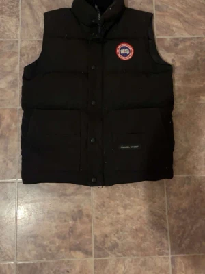 Svart dunväst från Canada Goose - Snygg svart dunväst från Canada Goose med hög krage och två stora fickor framtill. Västen har klassisk logopatch på bröstet och stängs med tryckknappar. Perfekt för lager-på-lager och håller dig varm under kyliga dagar.