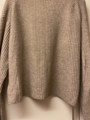 Beige ribbstickad tröja - Säljer en beige ribbstickad tröja med lång ärm och rak passform. Tröjan har en mjuk struktur och är tillverkad i ett tjockare stickat material, perfekt för kyligare dagar. Enkel och stilren design utan synliga loggor eller detaljer. Färgen är blandning av beige och en annan häftig färg  