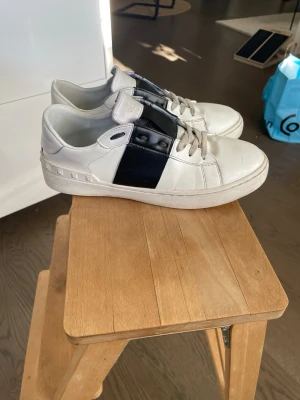 Valentino open - Snygga vita sneakers från Valentino med svart detalj på sidan och coola nitar längs hälen. Skorna har klassisk låg profil, vita snören och är tillverkade i läder. Perfekta för dig som vill ha en stilren men edgy look.