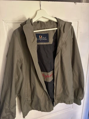 Grön vindjacka från Peak Performance - Snygg grå vindjacka från Peak Performance med huva och dragkedja framtill. Jackan har två sidofickor med dragkedja och innerficka. Materialet är tunt och vindtätt, perfekt för blåsiga dagar. Peak Performance-logga broderad på insidan. Passar M/L