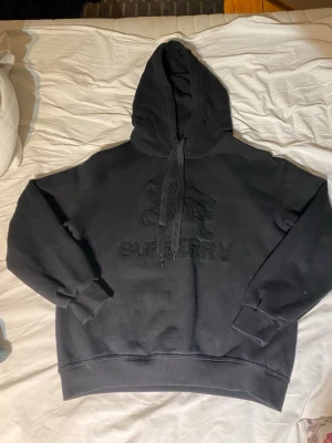 Svart hoodie från Burberry - Svart hoodie från Burberry med broderad logga och motiv på bröstet. Klassisk modell med stor huva, dragsko och ribbade muddar vid ärmslut och midja. Tillverkad i mjukt material som känns skönt mot huden.