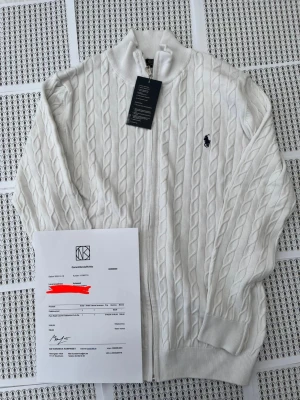 Vit kabelstickad ziptröja Polo Ralph Lauren - Kabelstickad vit ziptröja från Polo Ralph Lauren med klassisk marinblå logga broderad på bröstet. Tröjan har ribbade muddar, hög krage och dragkedja framtill. Perfekt för dig som gillar stilren och tidlös design.