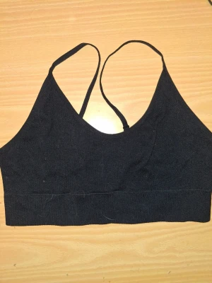 Svart sömlös bralette med smala band - En enkel och stilren svart bralette med smala axelband som korsas i ryggen. Bh:n är sömlös och utan bygel, vilket ger en mjuk och bekväm känsla. Materialet är stretchigt och följsamt, perfekt för en avslappnad look.