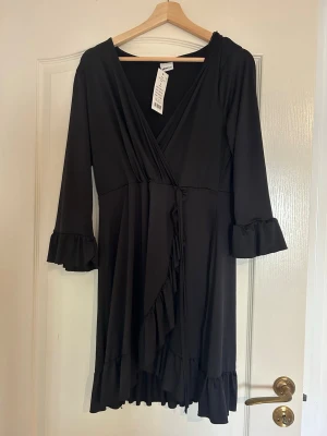 Svart omlottklänning Gina Tricot - Svart omlottklänning från Gina Tricot med volangdetaljer vid ärmslut och nederkant. Klänningen har v-ringning och trekvartsärmar, vilket ger en snygg och trendig look. Perfekt för dig som gillar stilrena och feminina plagg.